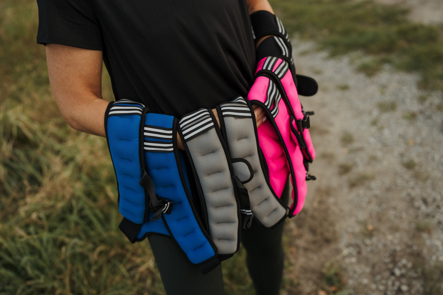 A person holding four weighted vests in front of their body. The vests are blue, grey, pink and black.